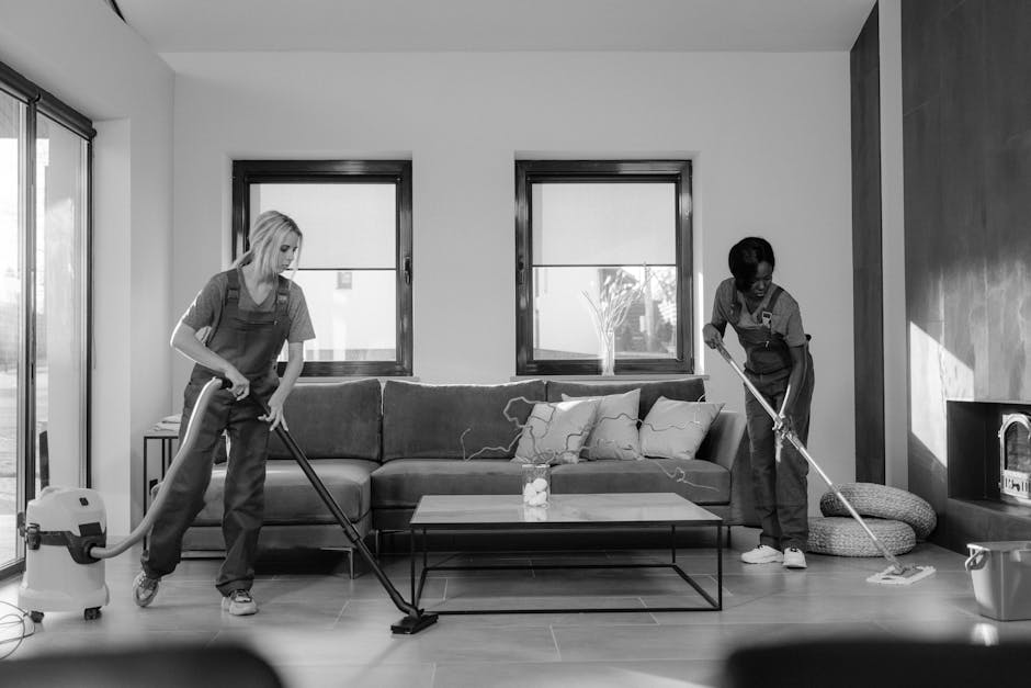 Three professional cleaners from London House Cleaning standing in a modern residential interior, wearing grey t-shirts and bright orange aprons. The female cleaner on the left holds a floor cleaning