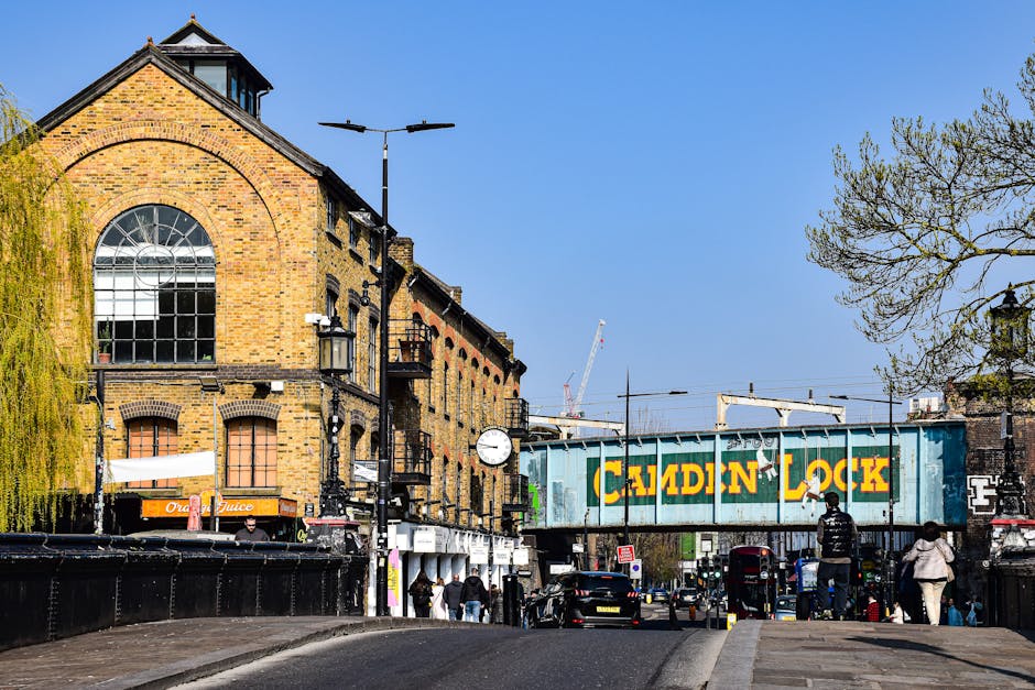 A photograph of Camden Lock in London showing a bustling street scene with a historic brick building featuring large arched windows on the left, a weathered blue railway bridge with bold yellow letter