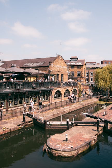 A photograph of Camden Lock in London showing a bustling street scene with a historic brick building featuring large arched windows on the left, a weathered blue railway bridge with bold yellow letter